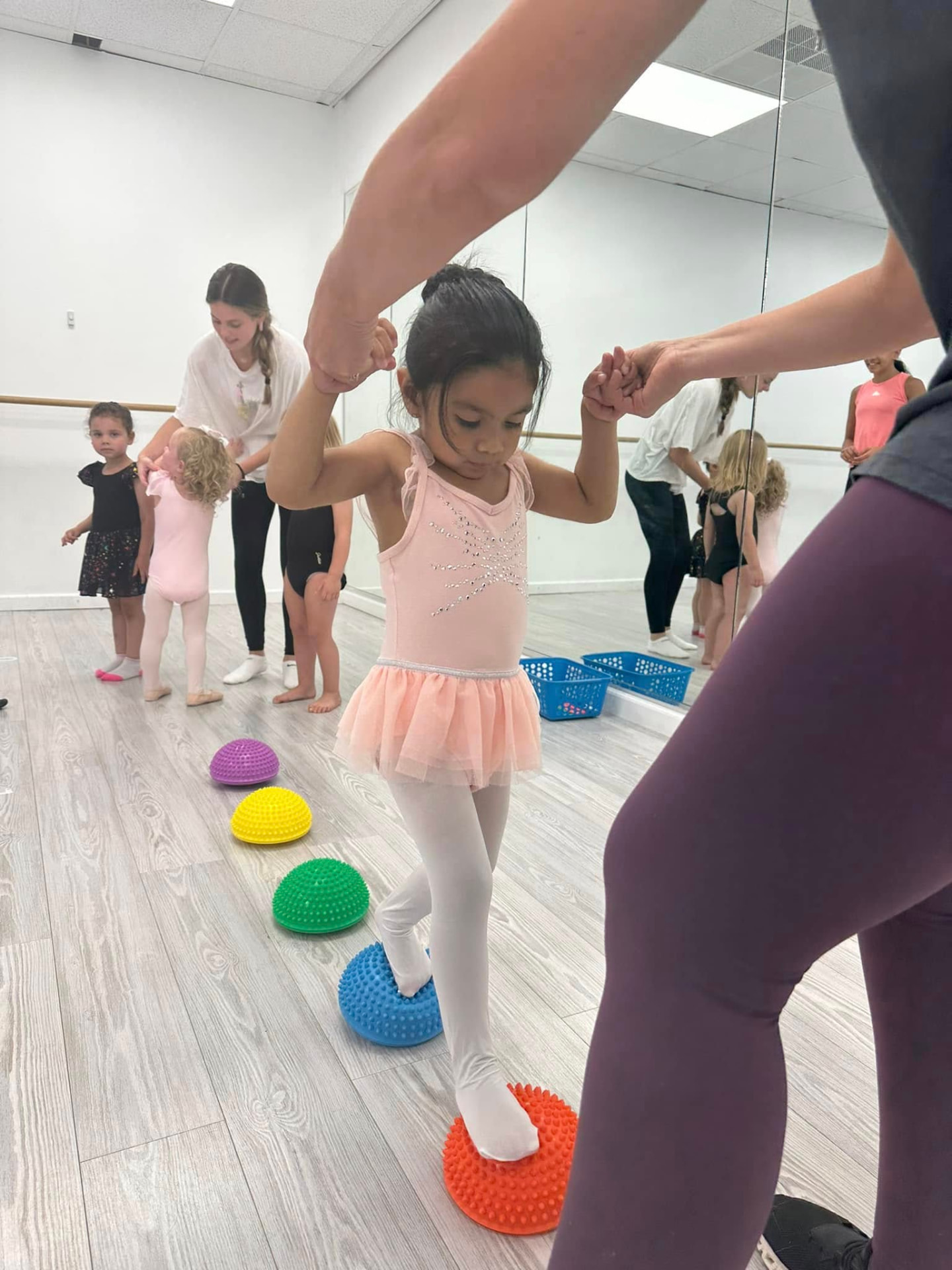 A young dancer balancing across colorful stepping stones with help from her teacher