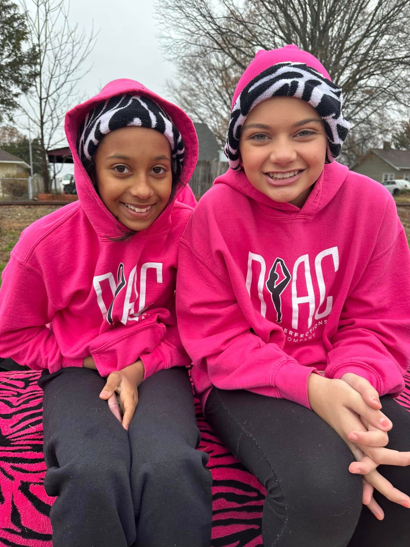 Two dancers in bright pink DPAC hoodies and zebra-print hats smiling outdoors