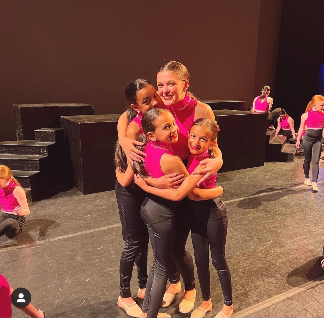 Dancers in pink costumes sharing a hug on stage after their performance