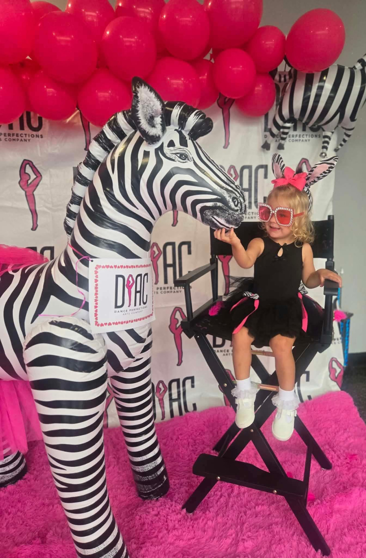 Young dancer in pink sunglasses and zebra ears petting a giant zebra in front of the DPAC backdrop and pink balloons