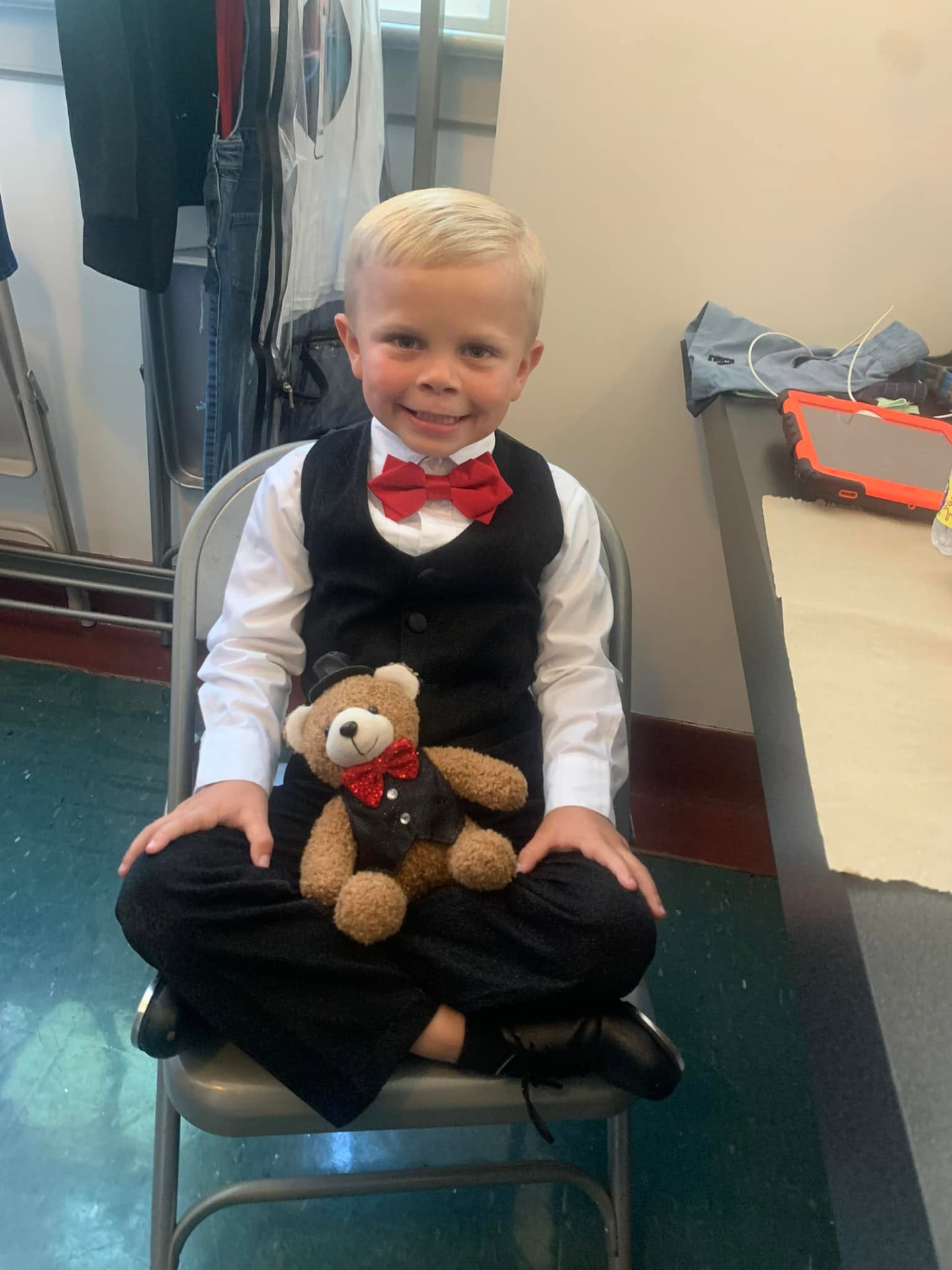 A young dancer in a tuxedo and red bow tie holding his matching teddy bear backstage